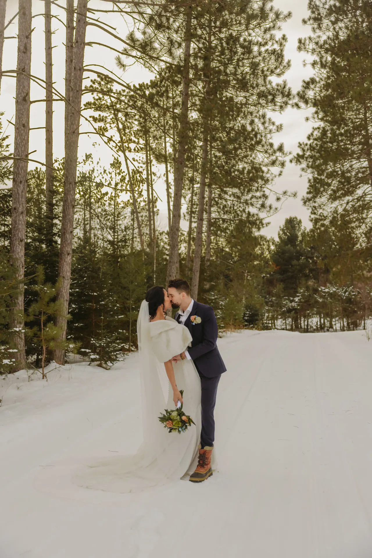 Bride in a long veil and groom in navy kissing on a snow-covered road lined with pines — Minneapolis & Twin Cities wedding photography by Emma Ziegler, Honey Deww Photography (Minnesota)