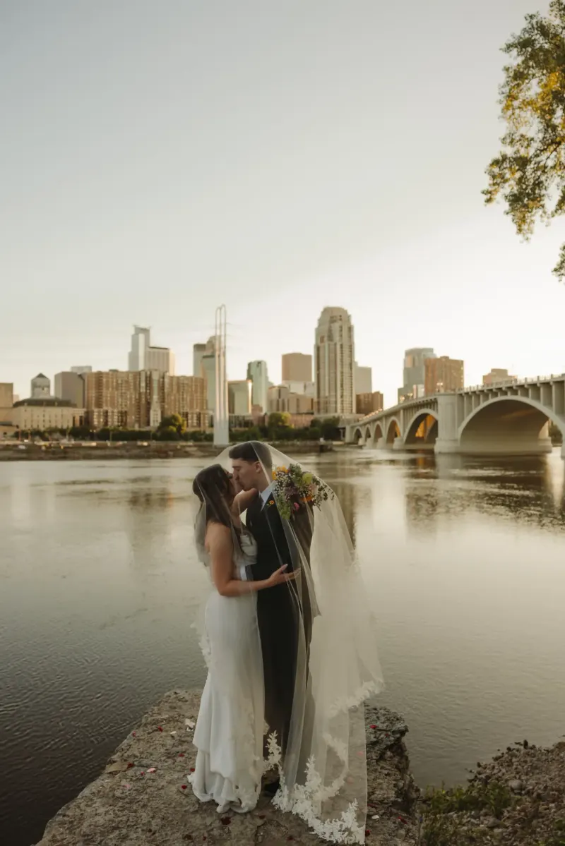 Two brides kissing beneath a veil on the Mississippi riverbank with the Minneapolis skyline and the Stone Arch Bridge behind them — the portrait stop a short walk from Mill City Museum, Nicollet Island Pavilion and The Machine Shop