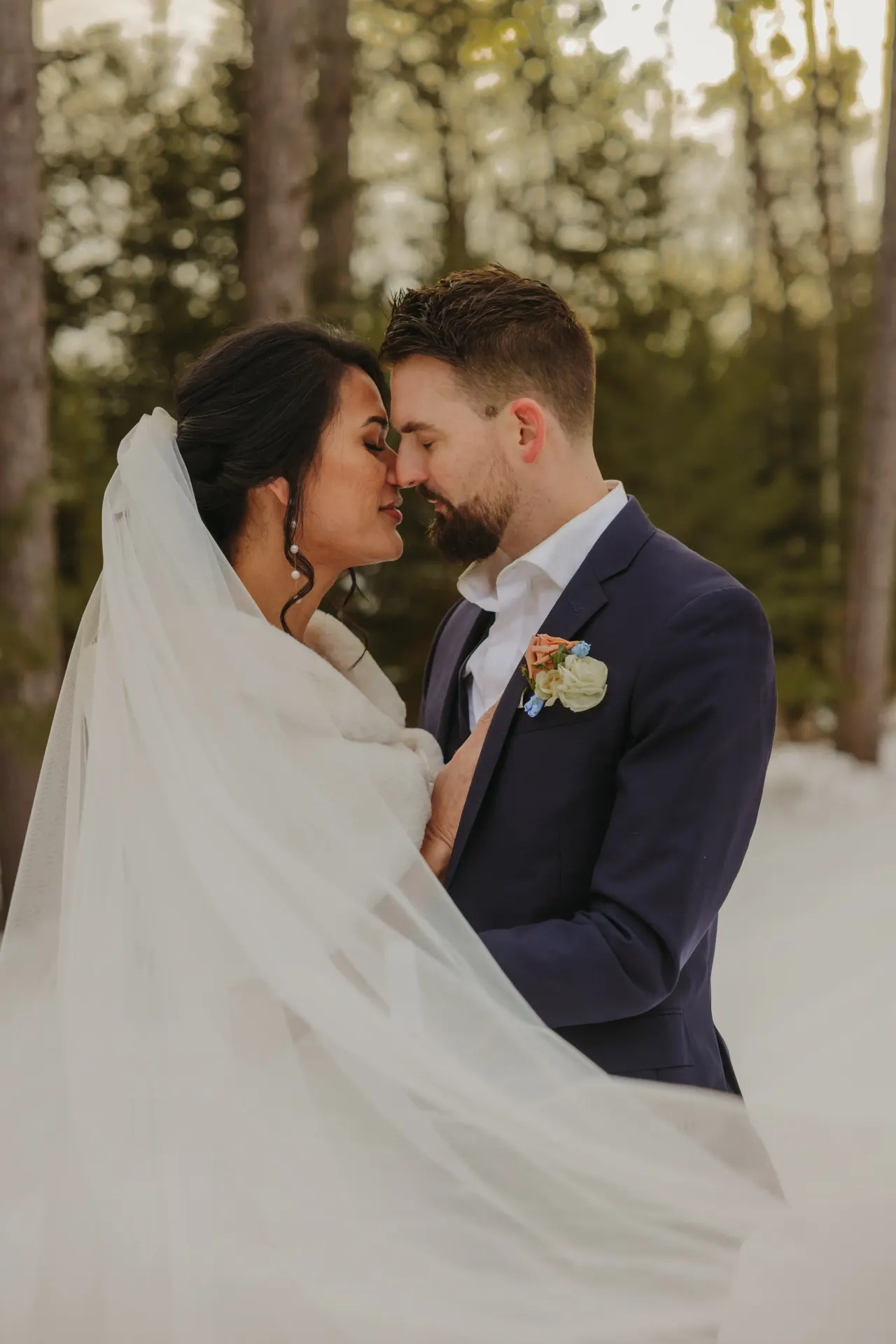 Bride and groom forehead to forehead in a snowy pine forest with her veil sweeping behind — Minneapolis & Twin Cities wedding photography by Emma Ziegler, Honey Deww Photography (Minnesota)