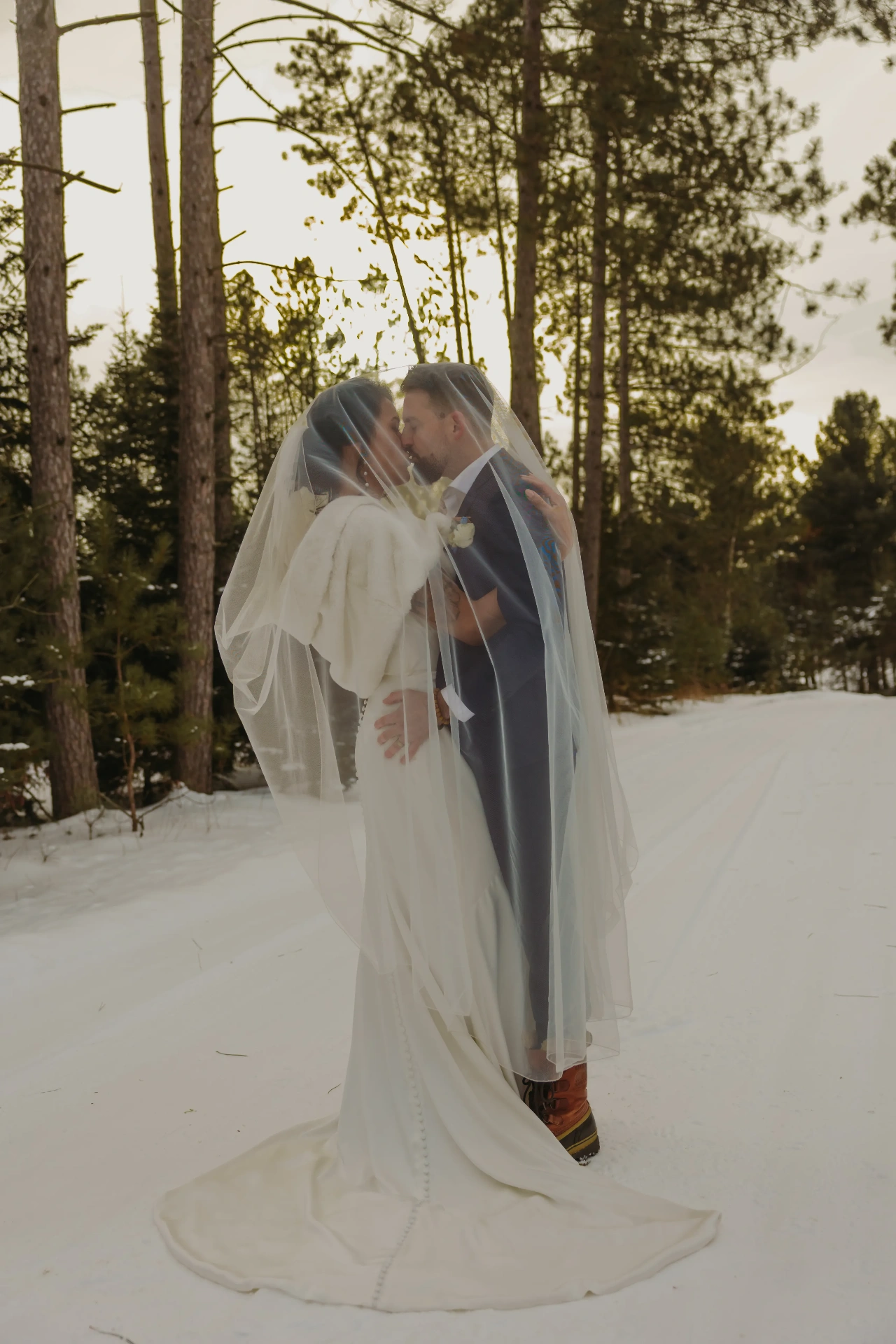 Bride and groom under veil kissing in snowy pine forest