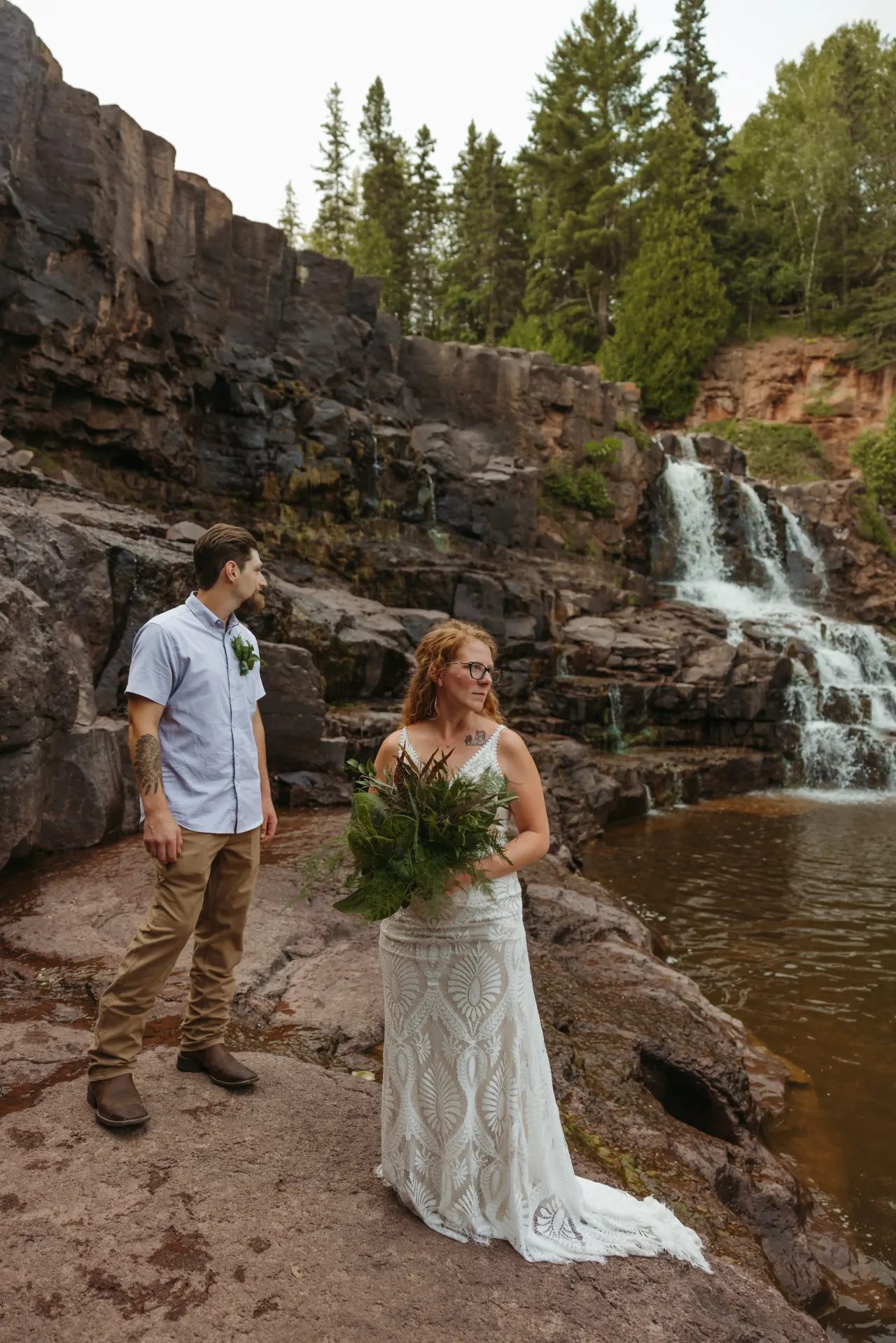 Bride holding a fern bouquet beside a groom on the rocks above a north-country waterfall — Minneapolis & Twin Cities wedding photography by Emma Ziegler, Honey Deww Photography (Minnesota)