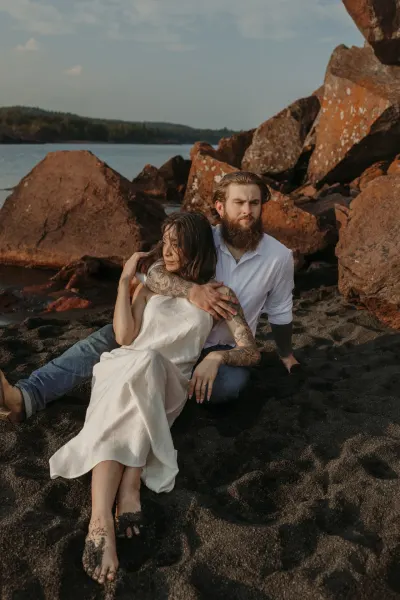 Couple sitting together on black sand beach with red rocks — Tonka Bay, Minnesota wedding photography by Emma Ziegler, Honey Deww Photography
