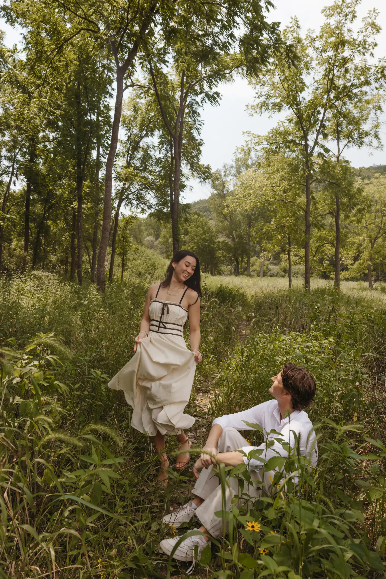 Bride gathering her skirt on a woodland path while her groom sits in the tall grass — Minneapolis & Twin Cities wedding photography by Emma Ziegler, Honey Deww Photography (Minnesota)