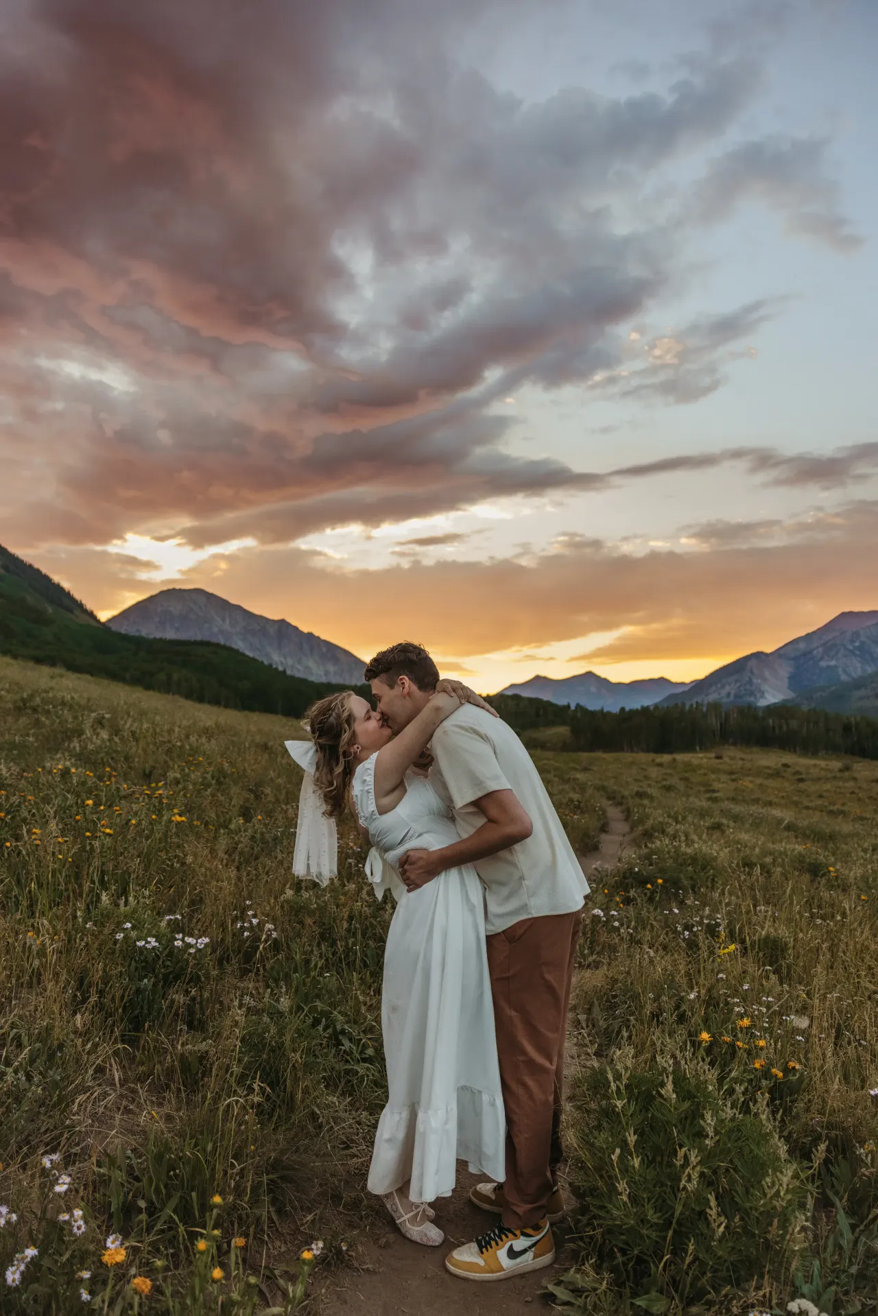 Bride and groom kissing on a meadow path beneath a pink and gold mountain sunset — Minneapolis & Twin Cities wedding photography by Emma Ziegler, Honey Deww Photography (Minnesota)