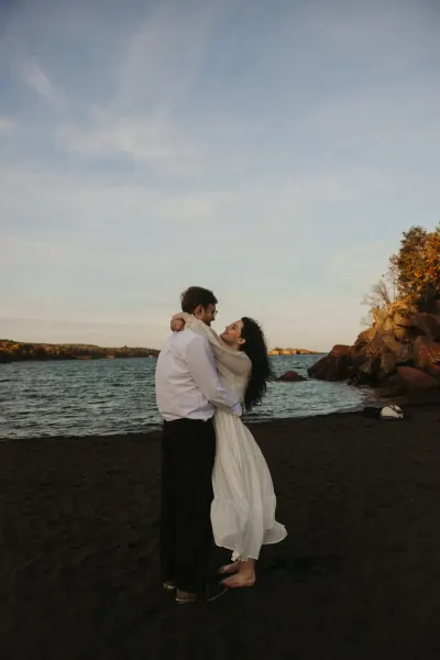 Couple embracing on dark sand beach with lake and rocks — Excelsior, Minnesota wedding photography by Emma Ziegler, Honey Deww Photography
