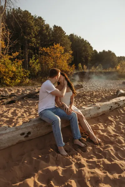 Couple kissing on driftwood log on sandy beach with autumn trees — Baldwin, Minnesota wedding photography by Emma Ziegler, Honey Deww Photography