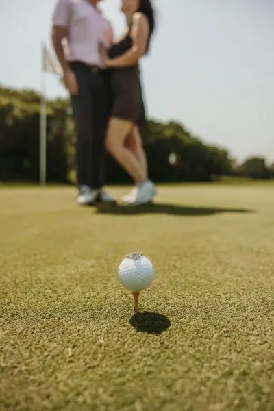 Engagement ring on golf ball with couple kissing in background — Deephaven, Minnesota wedding photography by Emma Ziegler, Honey Deww Photography