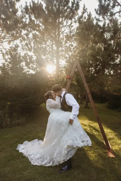 Groom lifting bride by wooden A-frame arch with sunburst through pines — Deephaven, Minnesota wedding photography by Emma Ziegler, Honey Deww Photography