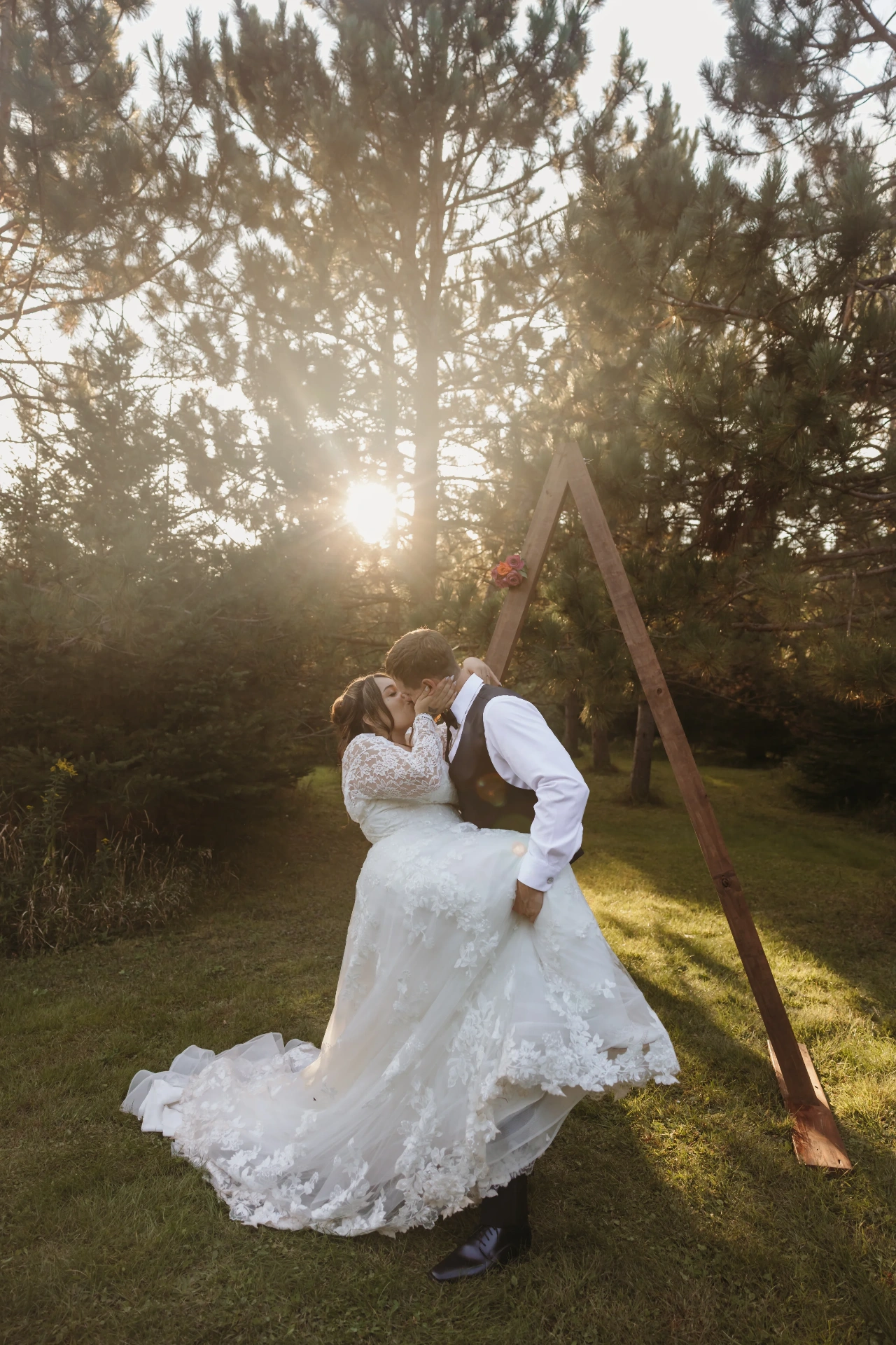 Groom lifting bride by wooden A-frame arch with sunburst through pines — Minneapolis & Twin Cities wedding photography by Emma Ziegler, Honeydew Photography (Minnesota)