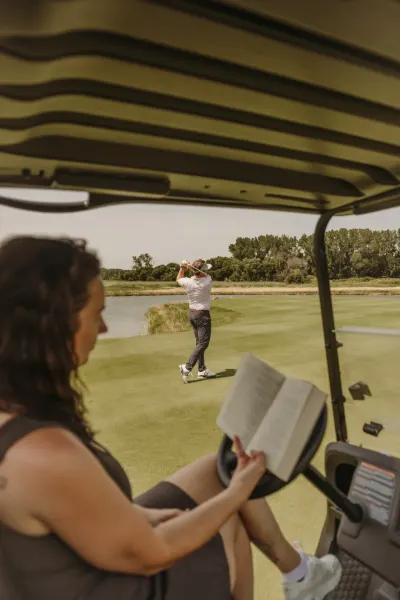 Woman reading in golf cart while man swings on course — St. Anthony, Minnesota wedding photography by Emma Ziegler, Honey Deww Photography