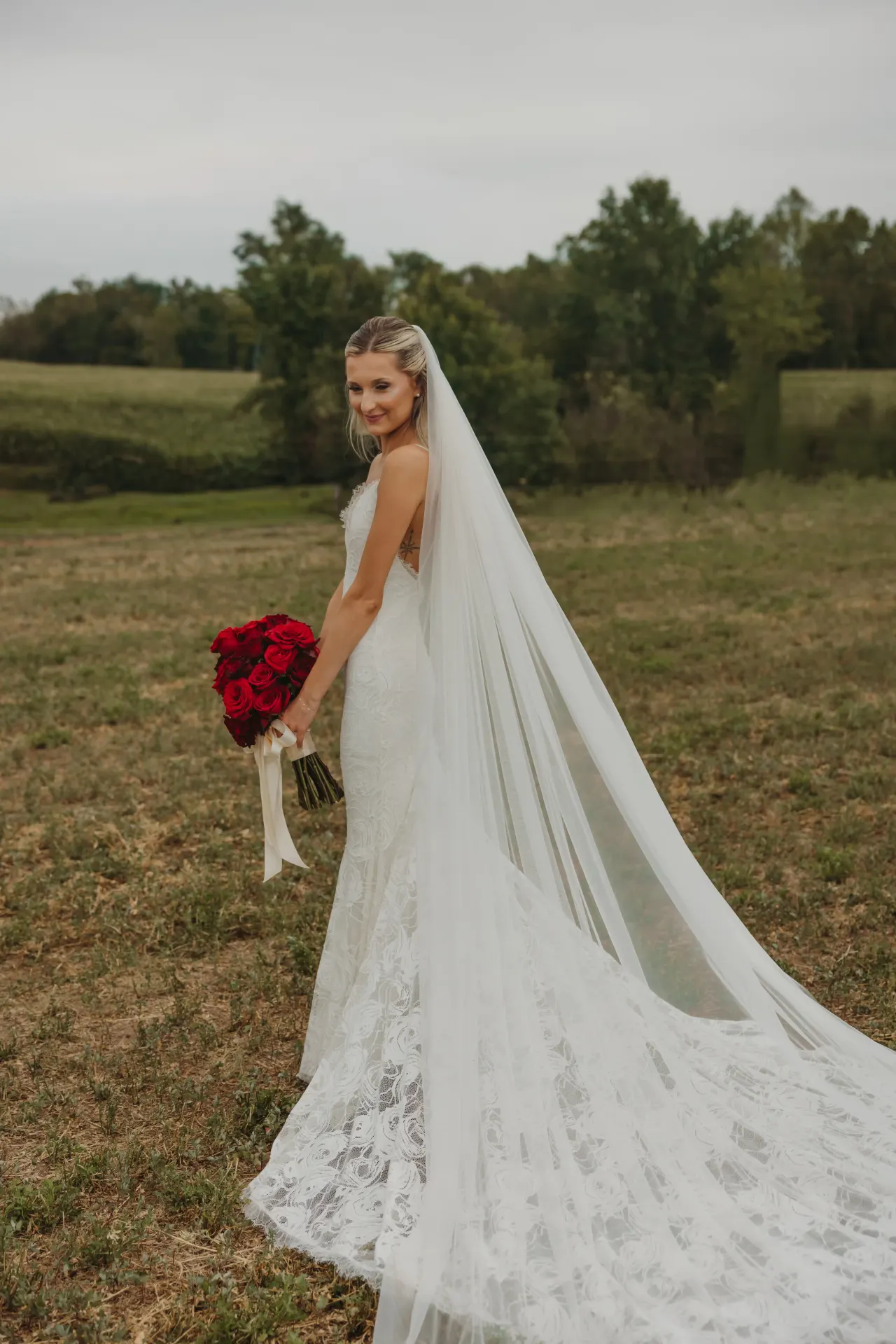 Bride from behind in a lace gown and cathedral veil, red roses in hand, in an open field — Minneapolis & Twin Cities wedding photography by Emma Ziegler, Honey Deww Photography (Minnesota)