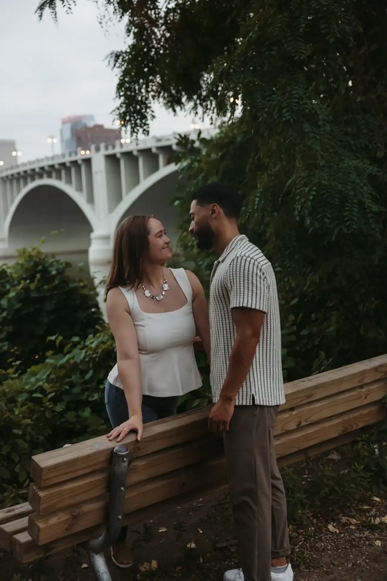 Couple leaning on a wooden rail with a river bridge and city skyline behind them — Minneapolis & Twin Cities wedding photography by Emma Ziegler, Honey Deww Photography (Minnesota)