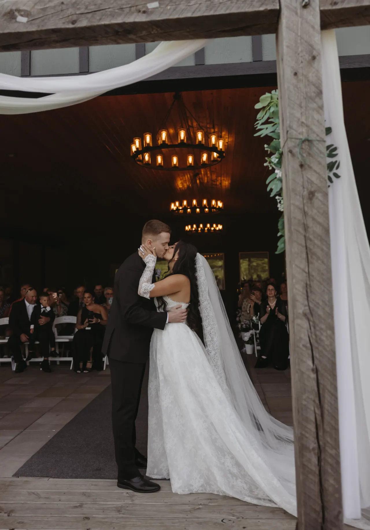 Bride and groom kissing at the end of the aisle beneath barn chandeliers — Minneapolis & Twin Cities wedding photography by Emma Ziegler, Honey Deww Photography (Minnesota)