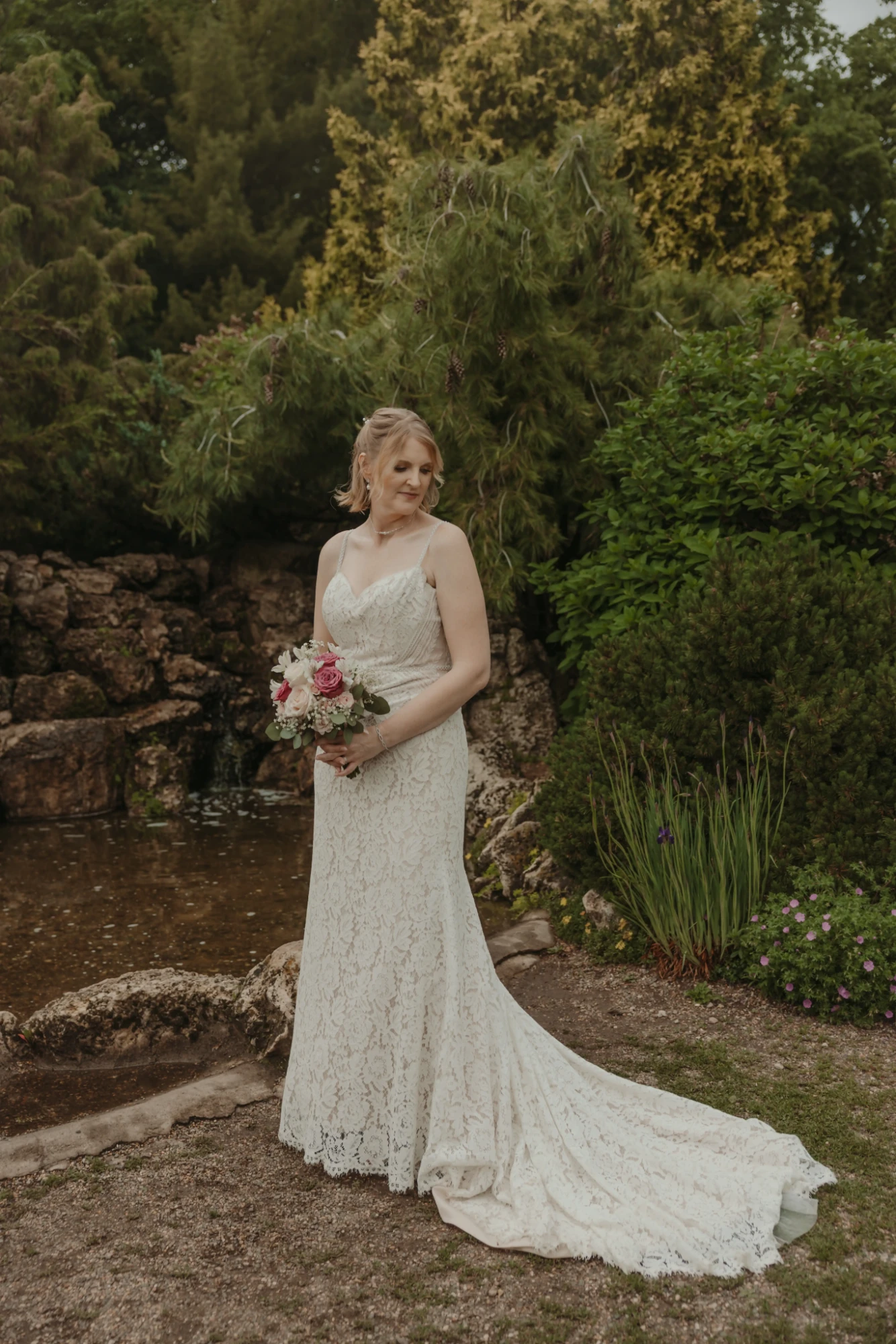Bride holding her pink and blush rose bouquet beside a rock-edged pond and evergreens at Lyndale Park Rose Garden