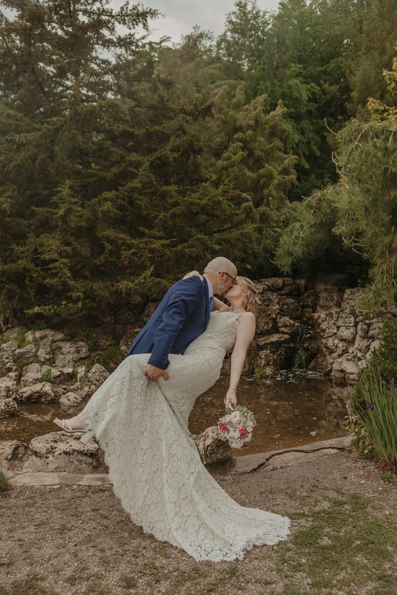Groom dipping the bride for a kiss beside the pond, her lace train spread on the path