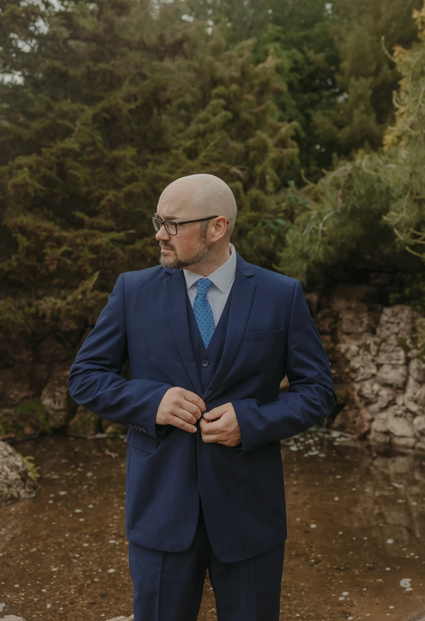 Groom in a navy three-piece suit buttoning his jacket by the pond