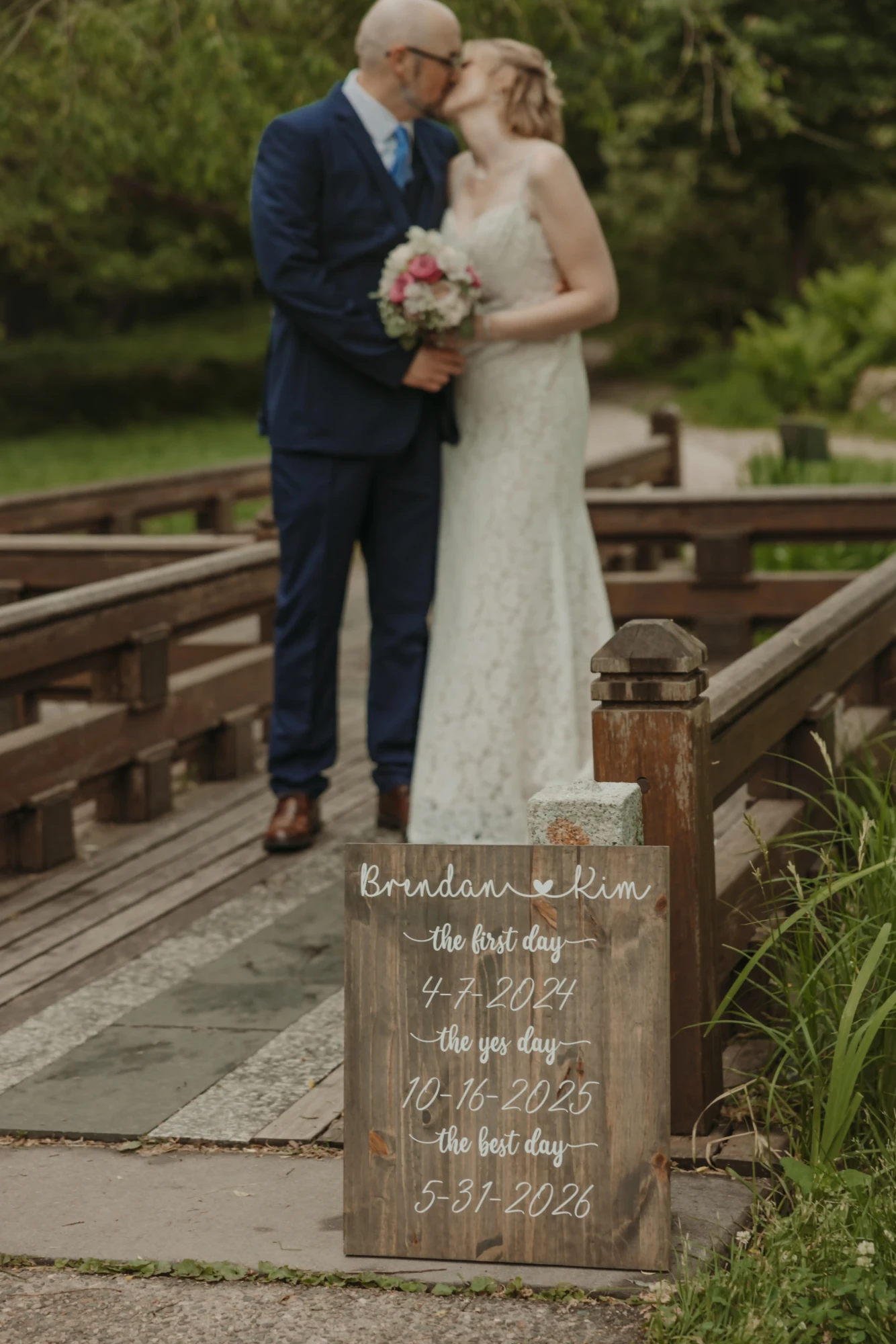 Couple kissing on the wooden footbridge behind their first day, yes day and best day sign