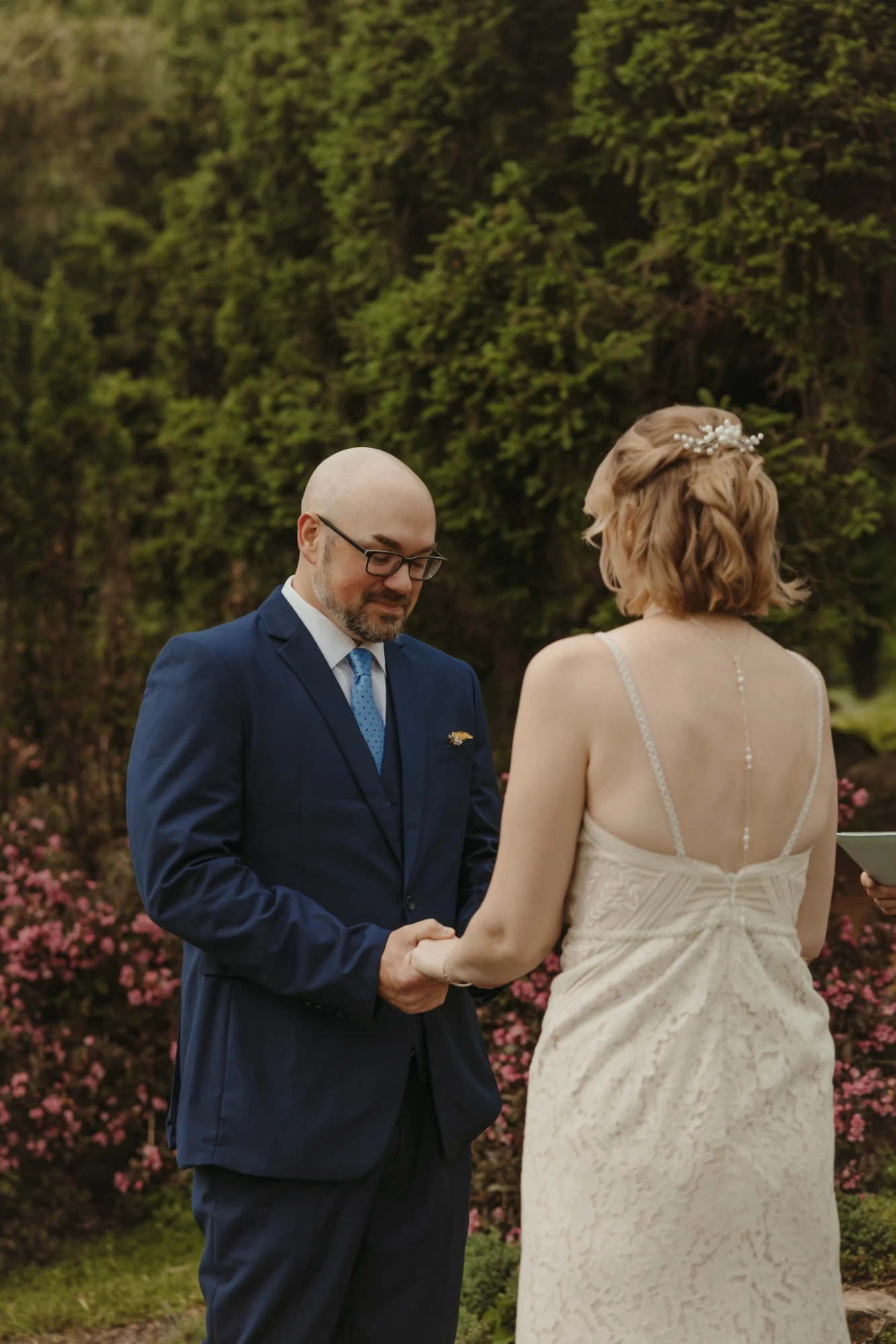 Couple holding hands during the vows in front of pink flowering shrubs