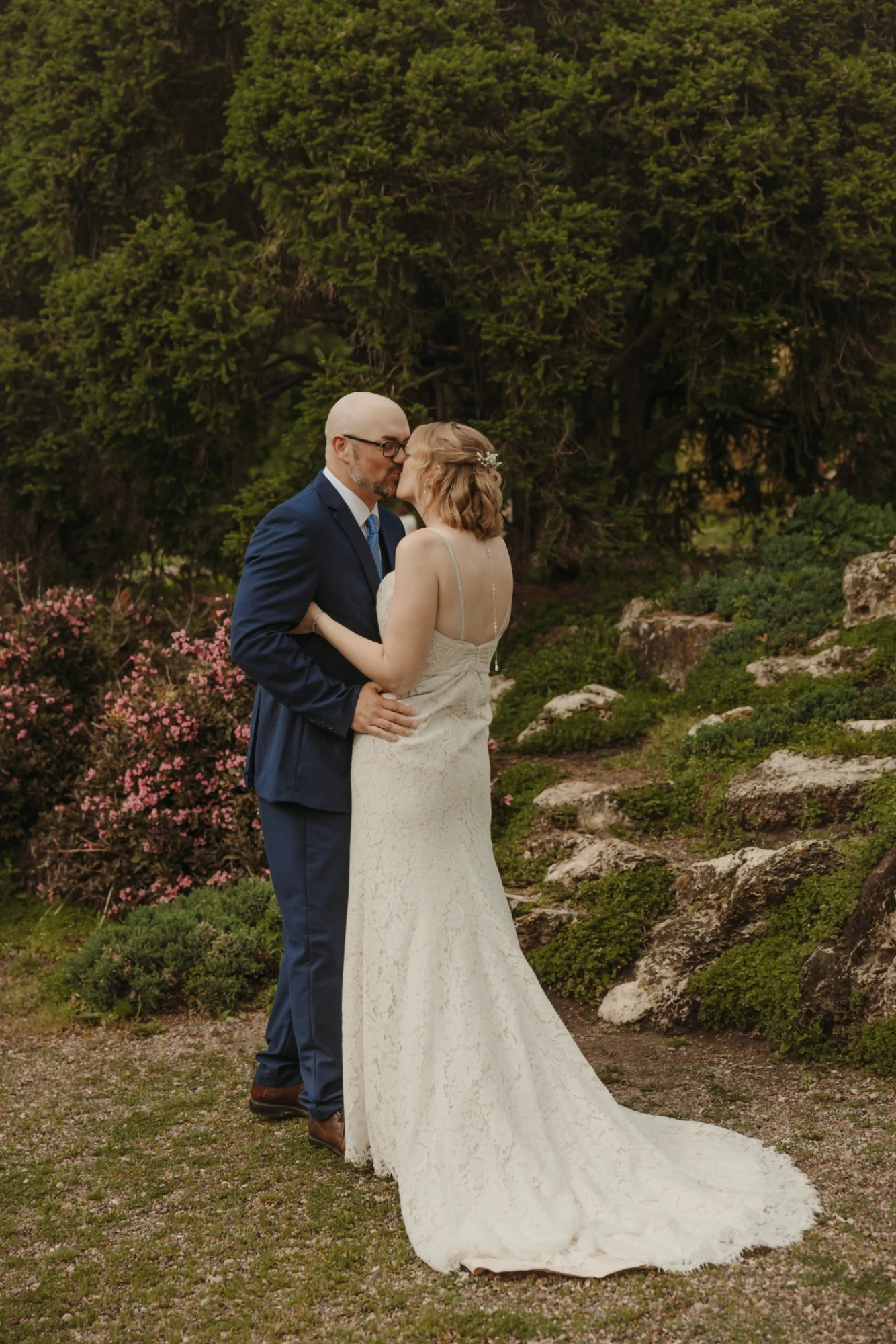 Couple kissing in front of pink shrubs, the gown's train behind them