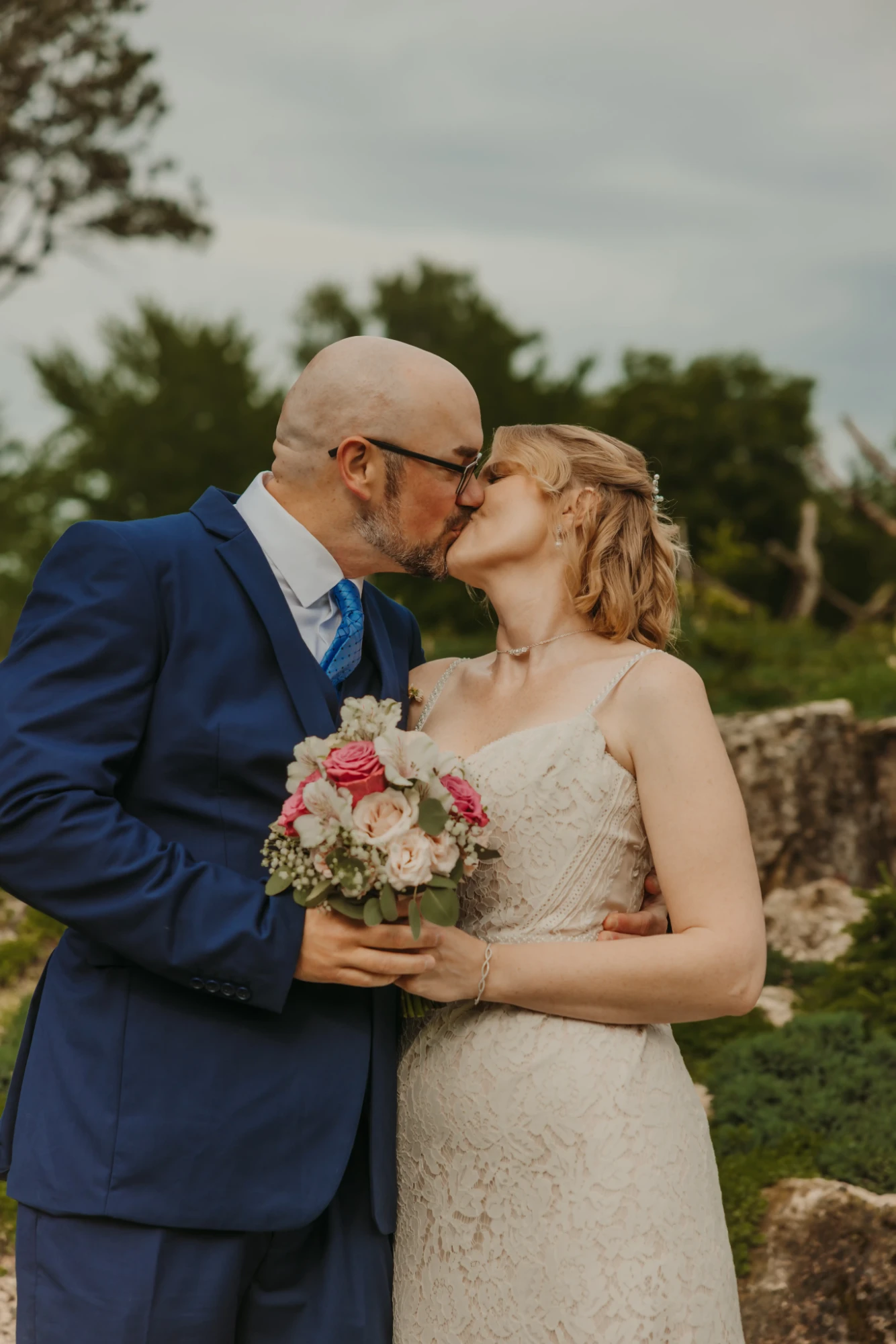 Couple kissing with the bouquet between them under an overcast sky